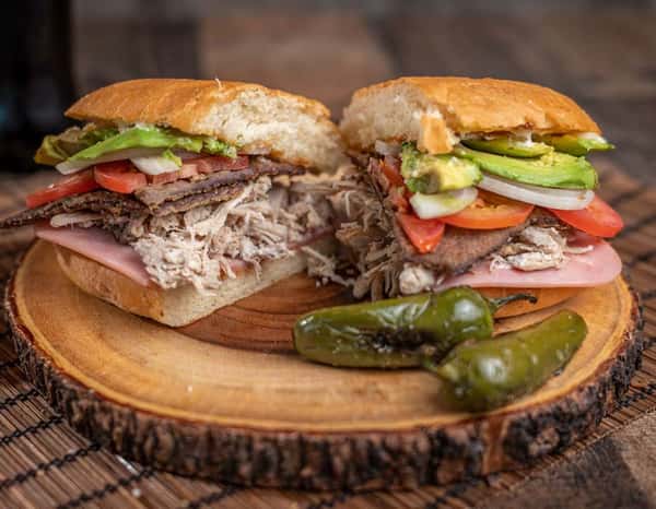 Mexican torta cut in half and served on a rustic wooden platter, layered with sliced ham, shredded pork, grilled beef, avocado, tomatoes, onions, and jalapeños on a soft bolillo roll — a stacked and savory sandwich perfect for traditional Mexican eateries or street food vendors.