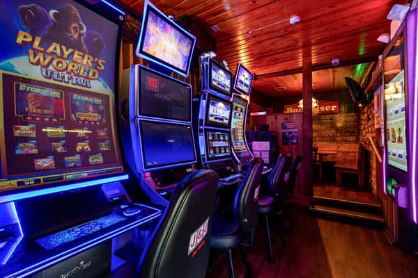 A cozy bar gaming area lined with brightly lit slot machines and black J&J Gaming chairs under a wooden ceiling.