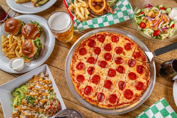Pepperoni pizza, mini burgers, fries, salads, and drinks on a wooden table in a lively casual dining setting.