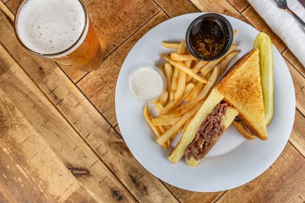French dip sandwich with roast beef on toasted bread, served with fries, au jus, horseradish sauce, a pickle, and a pint of beer.