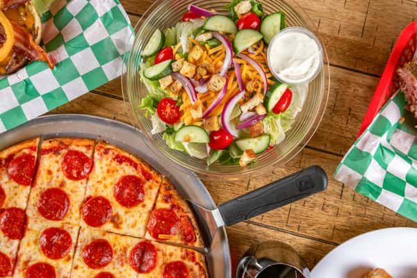 Pepperoni pizza, garden salad with ranch, bacon cheeseburger, and sandwich on a wooden table in a casual dining setting.