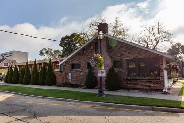 Street view of The Cottage restaurant, a brick building with patio area, green landscaping, and neon signs in the windows.