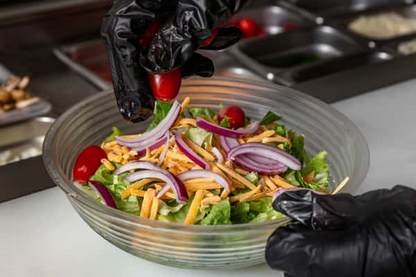Gloved hands preparing a fresh salad with lettuce, shredded cheddar, red onions, and grape tomatoes in a clear glass bowl.