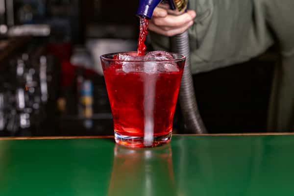 Red soda being dispensed into a glass filled with ice on a bar counter.
