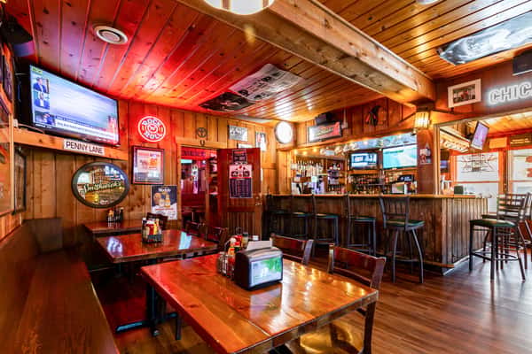Rustic indoor bar and dining area at The Cottage featuring wooden décor, TVs, neon signs, and a well-stocked bar with high stools.
