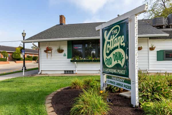 Front view of The Cottage restaurant with a vintage green sign reading "The Cottage Food/Drink, A Gathering Place" and garden landscaping.