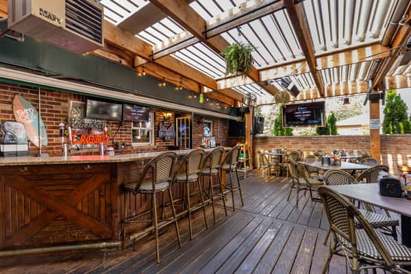 Covered outdoor patio with wooden floors, bar seating, and dining tables under a slatted pergola at The Cottage restaurant.