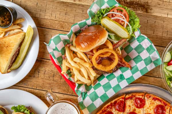 Bacon cheeseburger topped with onion rings, served with fries and fresh toppings in a red basket lined with green check paper.