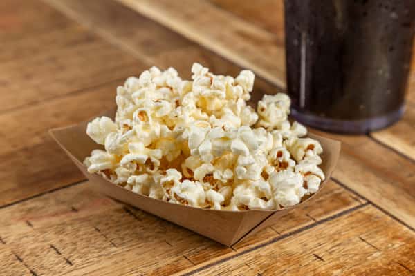 A small paper tray filled with freshly popped buttery popcorn, served on a rustic wooden table beside a cold drink—classic bar snack vibes.