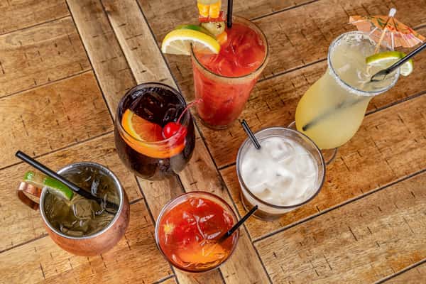 Assorted cocktails on a wooden table, including a margarita, Bloody Mary, Moscow mule, and drinks garnished with fruit.
