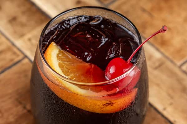 A glass of cola over ice garnished with a maraschino cherry and an orange slice on a wooden table.