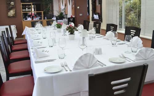Elegant indoor dining space with long white tablecloth-covered table, neatly set plates, wine glasses, and red-cushioned chairs.