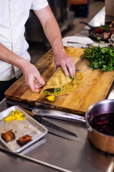 A chef in a white uniform is preparing a wrap on a wooden cutting board. The wrap is filled with greens and other ingredients, and the chef is carefully folding it. Fresh herbs and a pan with ingredients are visible nearby, along with a tray containing a few pieces of food.