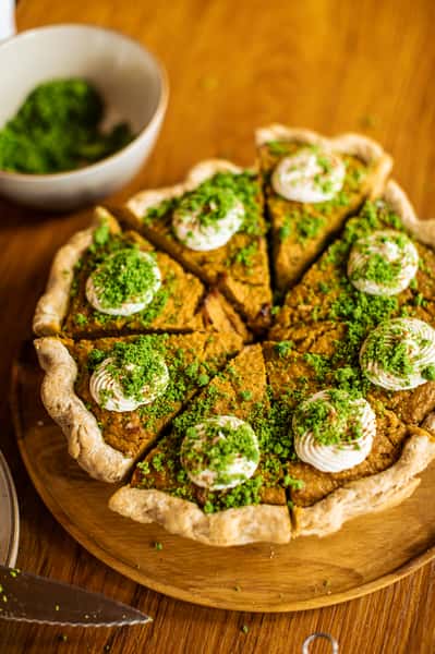 Pumpkin pie topped with whipped cream and green crumbles, sliced into wedges and served on a wooden board.
