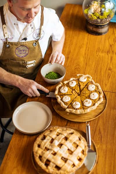 Chef in uniform slicing a pumpkin pie with whipped topping, a bowl of green garnish nearby, and an apple lattice pie on the table.