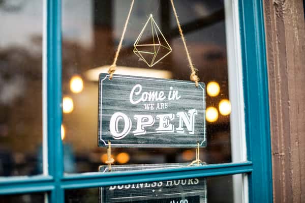 Hanging sign on a glass door that reads "Come in, we are OPEN" with warm lights blurred in the background.