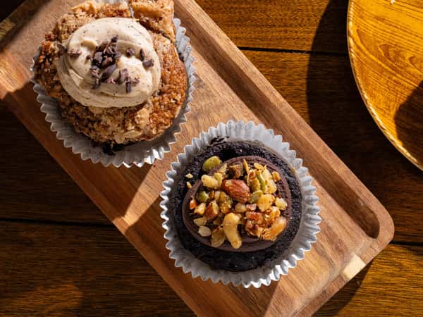 Tray with two muffins: one topped with frosting and cocoa nibs, the other a chocolate muffin topped with mixed nuts.