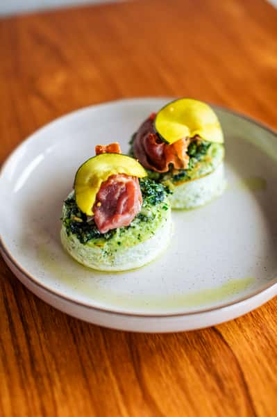 Two savory egg bites topped with greens, prosciutto, and thin zucchini slices served on a white plate with a drizzle of oil.