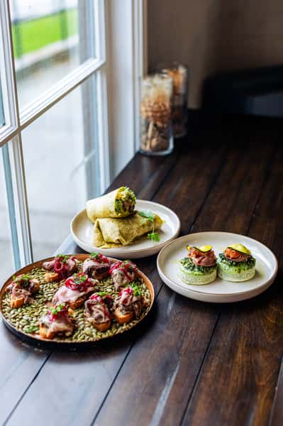 Plates of gourmet dishes by a window, including a wrap cut in half, small meat-topped appetizers on toast with seeds beneath, and two egg-based bites topped with meat and garnishes.