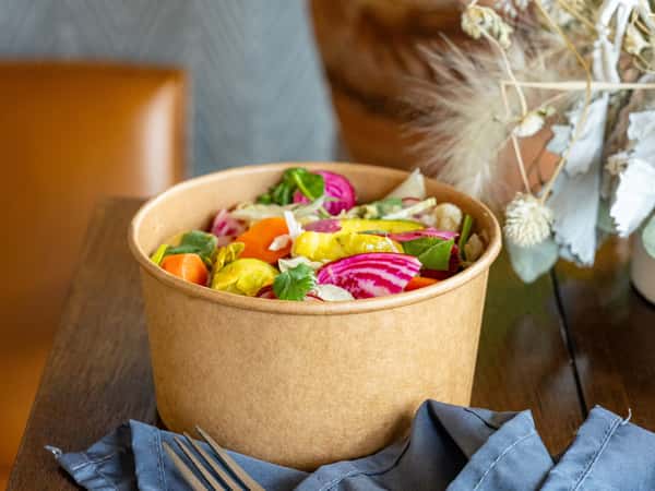 Colorful salad in a brown paper bowl with carrots, beets, squash, greens, and herbs, placed on a wooden table with forks.