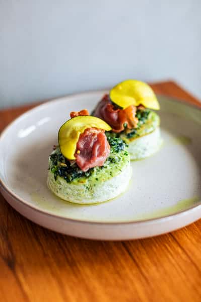 Two savory egg bites topped with greens, prosciutto, and thin zucchini slices served on a white plate with a drizzle of oil.
