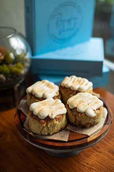 Four cinnamon rolls topped with swirls of cream cheese frosting on a wooden cake stand, with blue bakery boxes in the background.