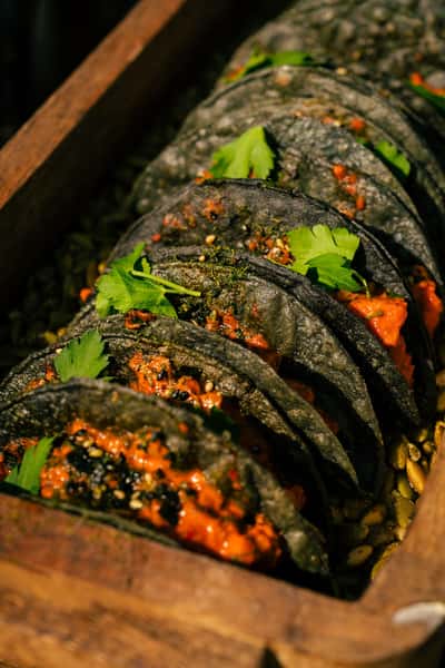 Charcoal black tacos filled with a rich red sauce and garnished with fresh herbs, neatly arranged in a wooden serving tray.