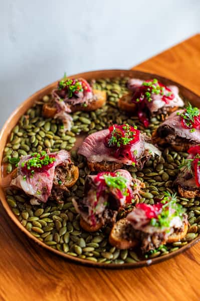 Crostini topped with tender sliced beef, pickled red onions, and fresh herbs served on a platter lined with pumpkin seeds.