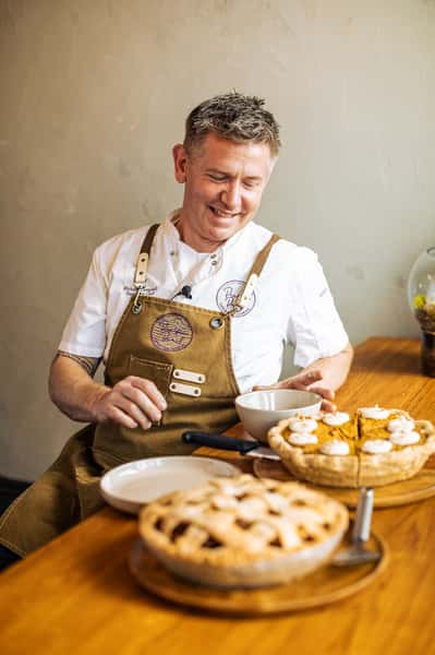 Chef smiling while preparing to serve slices of pumpkin pie with whipped cream, alongside a lattice-topped pie on the table.