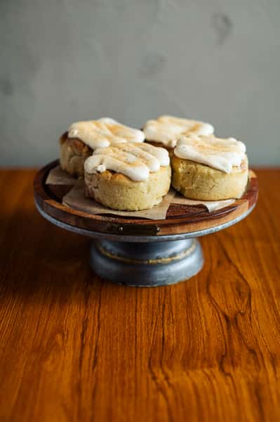 The image shows four cinnamon rolls topped with cream cheese frosting, displayed on a wooden pedestal stand lined with parchment paper.