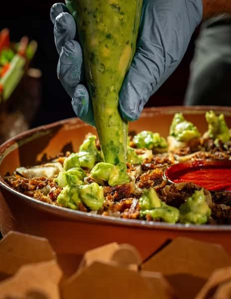Chef piping guacamole onto rolled tacos filled with meat, arranged around a bowl of red salsa on a large serving tray.