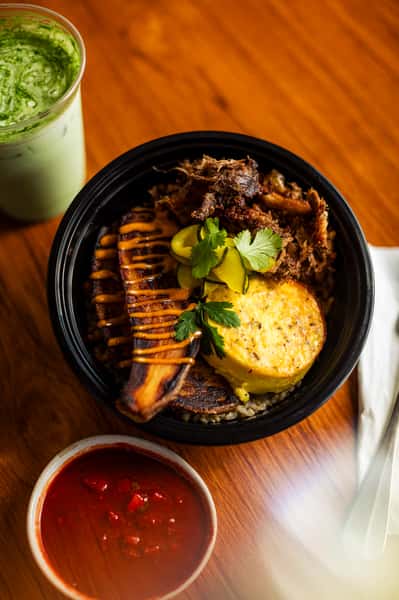 Savory rice bowl with roasted plantains, shredded meat, egg souffle, pickles, and cilantro, served with salsa and iced matcha.
