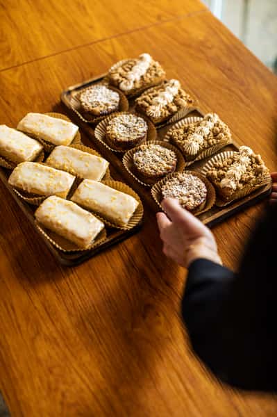 Trays of assorted pastries on a wooden table, including glazed rectangular cakes, crumb-topped rounds, and cream-topped bars.