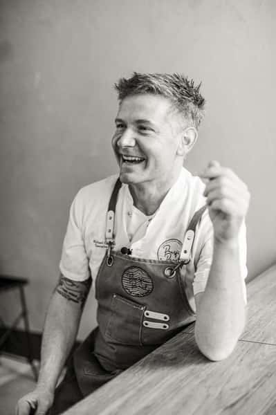 Smiling chef in a brown apron and white uniform sits at a wooden table, appearing cheerful and engaged.