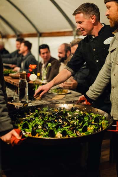 Chef serving a large pan of seafood paella with mussels and greens to guests seated at a long communal dining table.