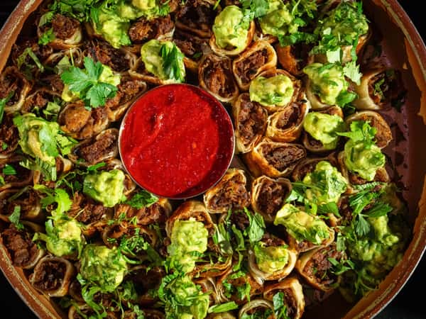 Platter of beef taquitos topped with guacamole and cilantro, served with a bowl of red salsa in the center.