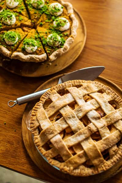 Pumpkin pie topped with whipped cream and green garnish beside a golden lattice apple pie on wooden serving boards.