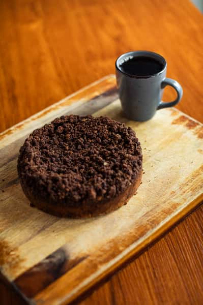 Chocolate crumb cake on a wooden board paired with a dark ceramic mug filled with hot black coffee.