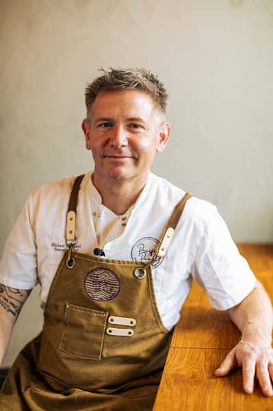 Chef in a white uniform and brown apron sitting at a wooden table, looking at the camera with a calm, confident expression.