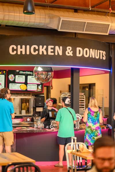 Customers ordering at a vibrant "Chicken & Donuts" counter inside a modern food hall, featuring a neon-lit disco theme with a shiny mirror ball, colorful signage, and a casual, fun atmosphere perfect for fried comfort food lovers.