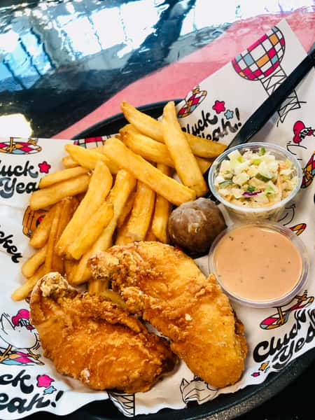 Crispy fried chicken tenders served with thick-cut fries, creamy coleslaw, a glazed donut hole, and a side of zesty dipping sauce, all plated on branded paper with playful "Cluck Yeah!" graphics — a bold and indulgent American comfort food combo.