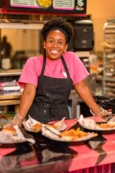 Smiling staff member in a pink shirt and black apron standing behind the counter at a chicken and donuts restaurant, with trays of fried chicken meals ready to serve — a welcoming and vibrant fast-casual dining experience.