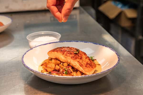 chef plating a seared salmon dish over vegetables with sauce in a restaurant kitchen