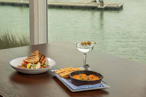 waterfront dining table with baked seafood dip, crackers, burrata salad, and martini overlooking the marina