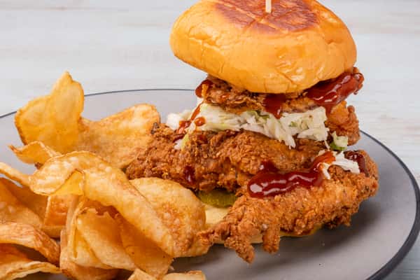 Fried chicken sandwich topped with slaw and sauce on a toasted bun, served with potato chips on a plate at a restaurant