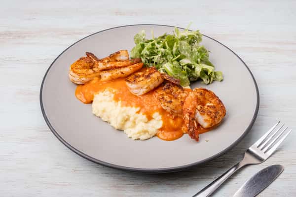 Grilled shrimp and creamy grits topped with a spicy sauce, served with a side salad on a gray plate at a restaurant