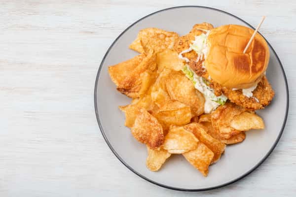 Crispy fried fish sandwich with lettuce on a bun, served with potato chips on a gray plate at a restaurant