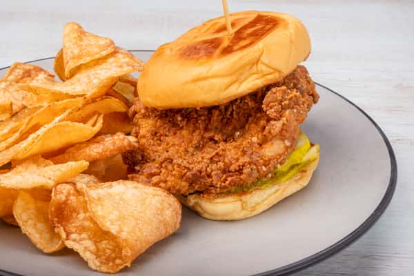 Fried chicken sandwich with pickles on a toasted bun, served with potato chips on a gray plate at a restaurant