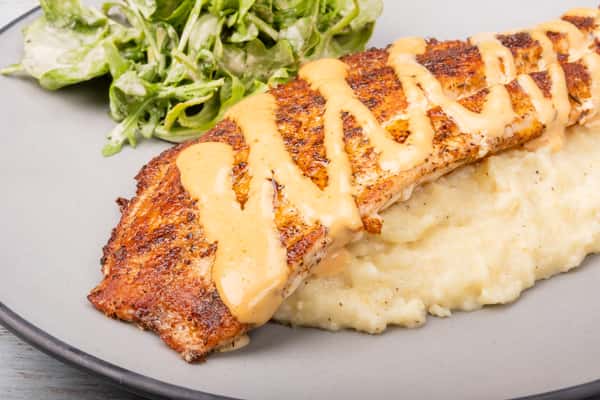 Blackened red snapper drizzled with a creamy sauce, served over mashed potatoes with a side salad on a gray plate at a restaurant