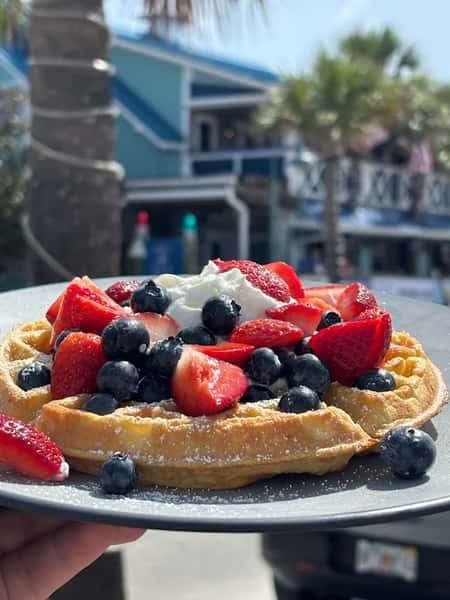 Belgian waffle topped with fresh strawberries, blueberries, whipped cream, and powdered sugar served outdoors on a sunny patio